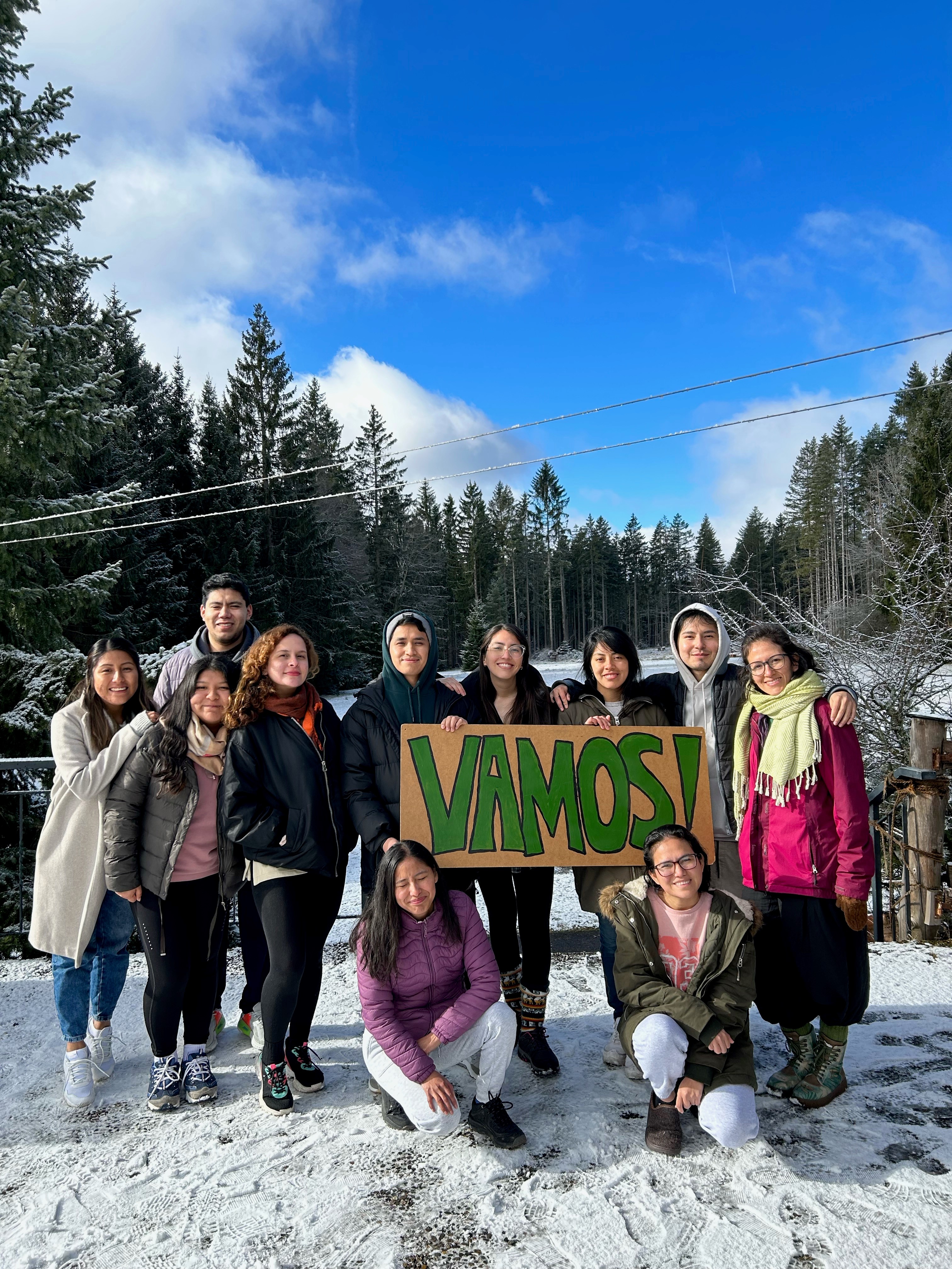 Gruppenfote der Vamos Freiwilligen mit Betreuern im Schnee. Die Freiwilligen halten ein Pappkarton Schild hoch, auf dem in grün VAMOS! steht. Im Hintergrund blauer Himmel und beschneite Nadelbäume.